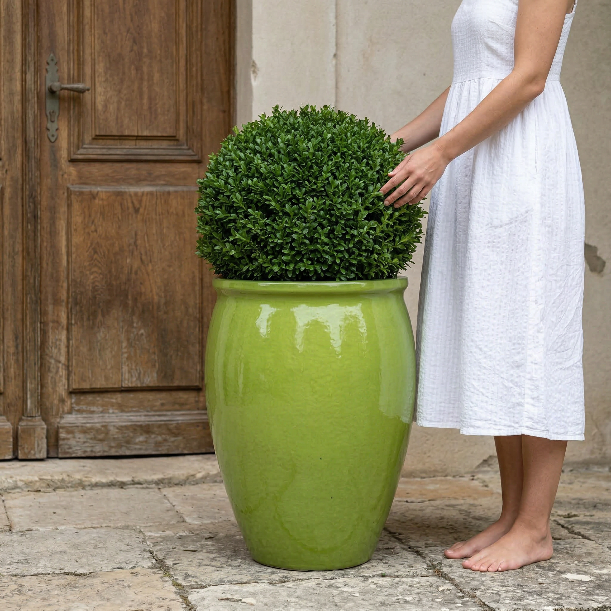 Keramický květináč BACCHUS - Olivově zelená s kulatým buxusem před starými dřevěnými dveřmi. Žena v bílých letních šatech se jemně dotýká rostliny.