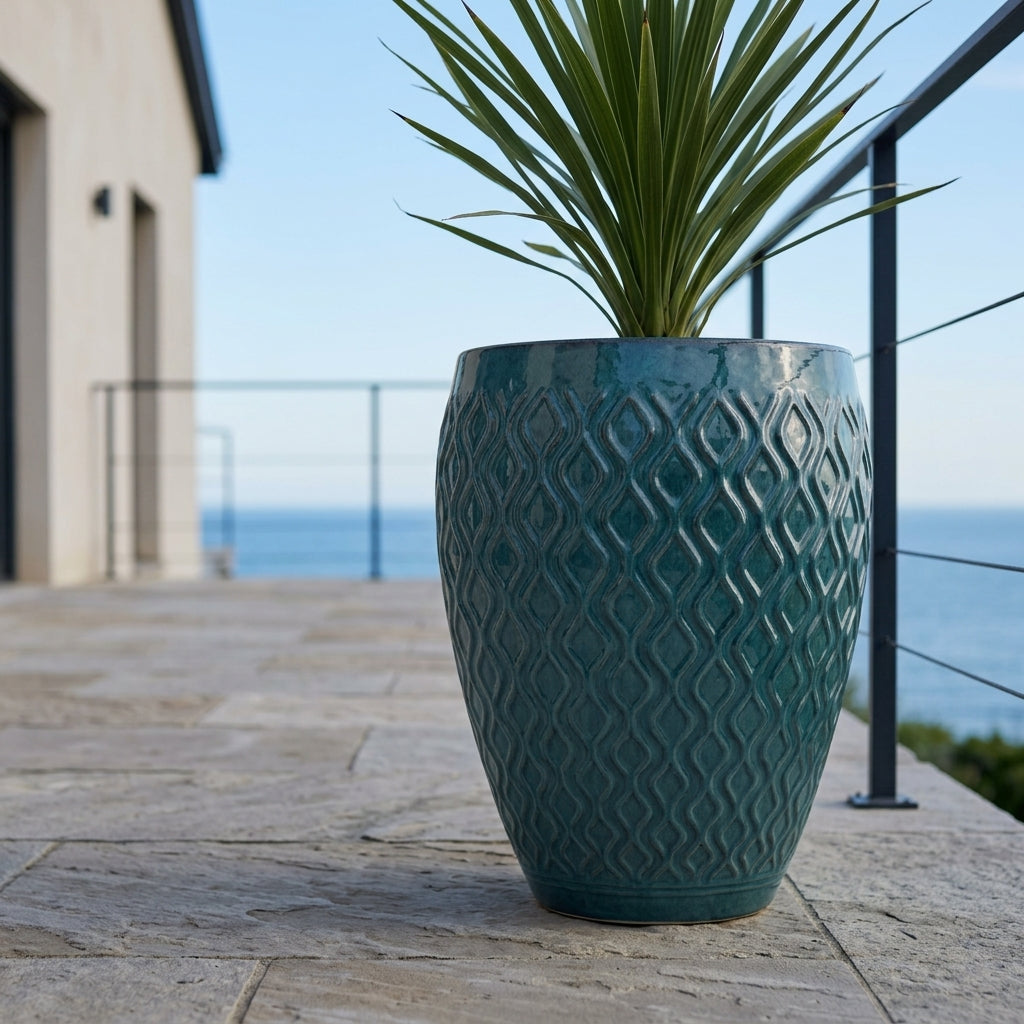Detailní záběr na petrolejově modrý keramický květináč Phenix s rostlinou Cordyline na kamenné terase s výhledem na oceán.
