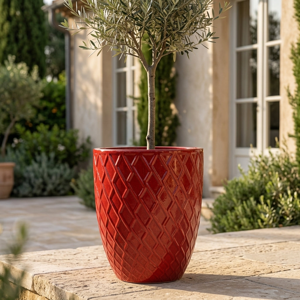Detailní pohled na lesklý keramický květináč Zeus - Císařská červená s ozdobným reliéfem. Elegantní velká nádoba tvaru urny s olivovníkem na prosluněné kamenné terase.