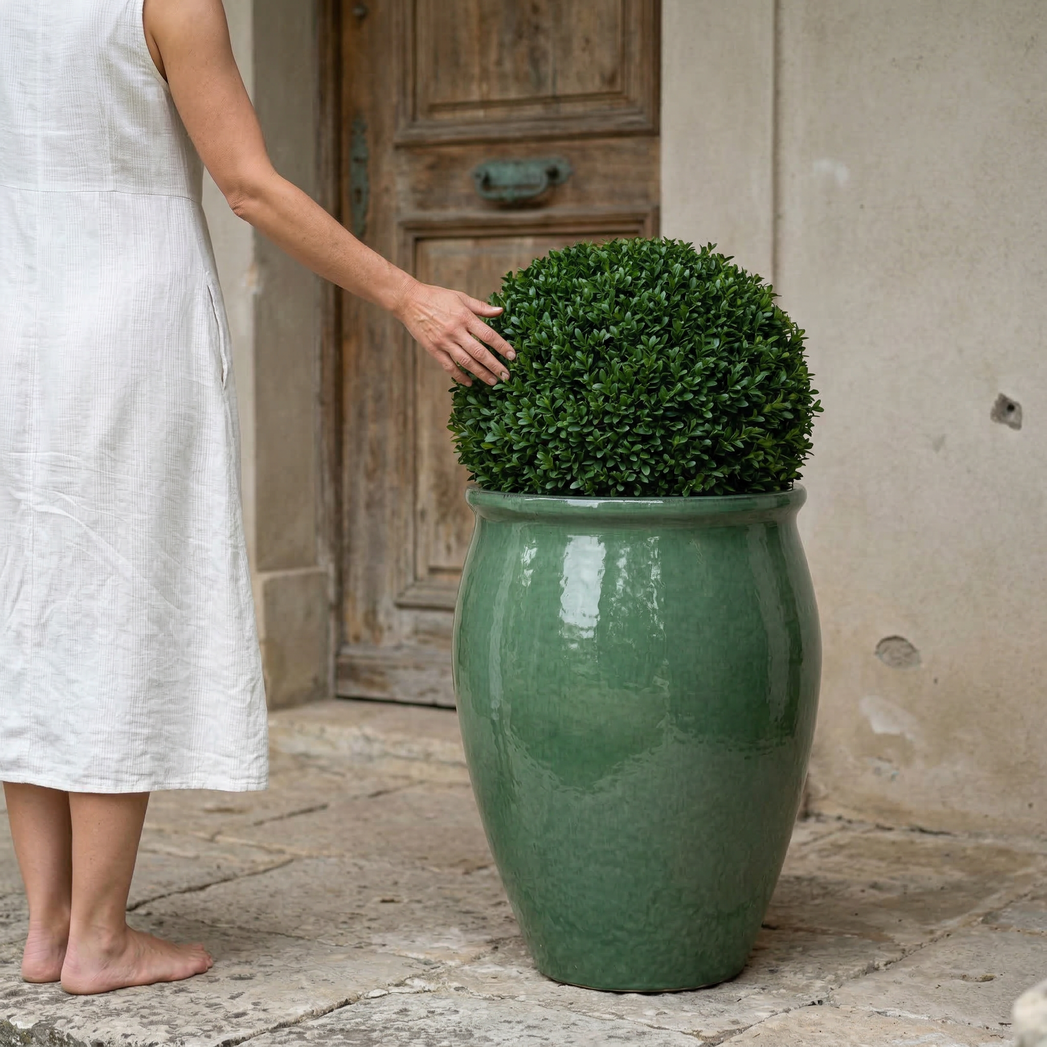 Keramický květináč BACCHUS - Nefrit s buxusem před starými dřevěnými dveřmi, u kterého žena v bílých letních šatech pečuje o rostlinu.