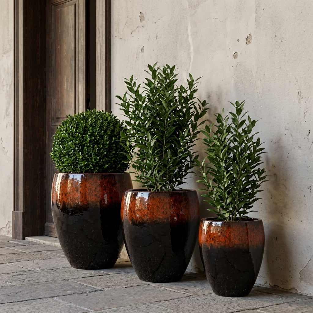 Detail na velký keramický květináč Poseidon - Černo oranžová s kulatým buxusem před starými dřevěnými dveřmi. Žena v bílých šatech se jemně dotýká rostliny.