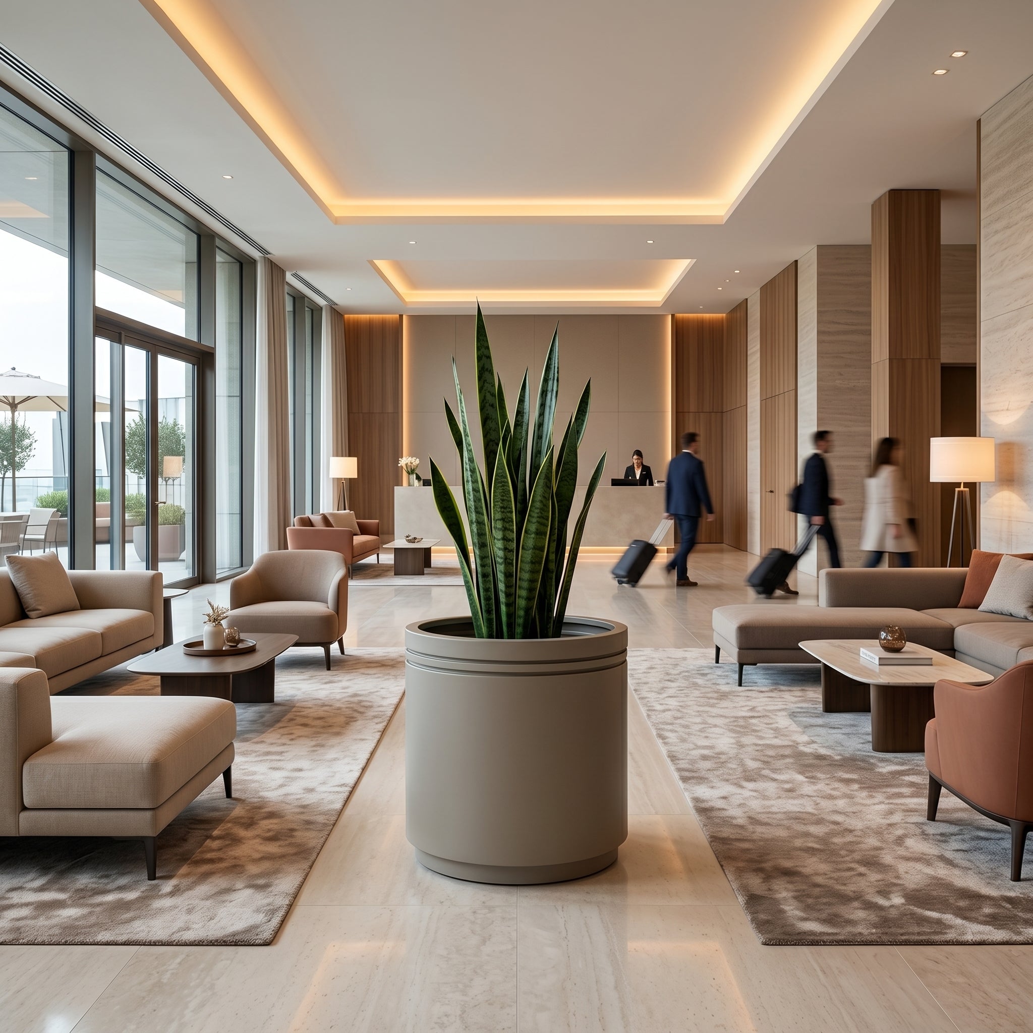 Velký pozinkovaný ocelový květináč INFINITY ROUND - Šampaňská - XL (60 x 60 x 60 cm) s rostlinou Sansevieria uprostřed moderního lobby luxusního hotelu s procházejícími hosty v pozadí.